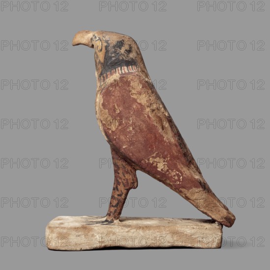 Figurine of the falcon Sokaris, guardian of the necropolis; Horus mythology; Sokaris mythology. A falcon stands on a thick, rectangular base, resting its tail on the back edge of the base. The bird's head is slightly raised. The beak is long, curved; the body is long, with wings slightly plastically defined in their upper part. The lower part of the wings merges with the tail. The head is black on the sides and back. The front of the head is yellow with a black, diagonal check. The eye is marked with white and black paint. A white stripe around the neck. The wings are black and red. Bird legs with red and black lines. Late Period–Ptolemaic Period, 664–30 BC, Egypt. Sculpture; height 24.3 cm, width 20.5 cm., A sculpted bird figure standing on a rectangular base, depicted in profile with a curved beak and a streamlined body; the surface shows worn painted areas including reddish-brown on the body, beige and tan patches where the paint has flaked, black detailing around the head and neck,...