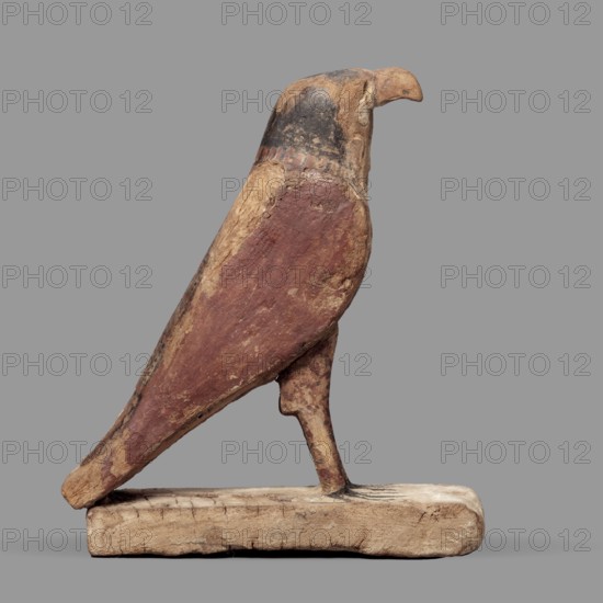 Figurine of the falcon Sokaris, guardian of the necropolis; creator, Horus mythology. A falcon stands on a thick rectangular base, resting its tail on the back edge. The bird's head is slightly raised; the beak long and curved; the body long with wings slightly plastically defined in their upper part and the lower parts merging with the tail. The head is black on the sides and back, the front of the head yellow with a black diagonal check; the eye marked with white and black paint; a white stripe around the neck; wings black and red; bird legs with red and black lines. Sokaris mythology. Date: 664–30 BC, Late Period–Ptolemaic Period, Egypt. Sculpture. Height 24.3 cm, width 20.5 cm., A carved bird figure perched on a rectangular base, shown in profile, with a rounded head and hooked beak, a tapered body and a single visible leg attached to the base; the surface appears worn and textured with visible cracks and areas of abrasion. The colors visible include shades of brown, reddish-brown,...