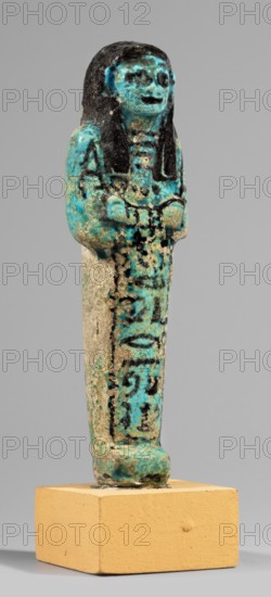 Shabti of the Lady of Renu, Mummy-shaped figurine made of blue faience with dark details. Arms crossed on the chest, right over left, holding hoes in both hands. Wears a three-part smooth wig finished with borders; round face, large eyes, wide nose, prominent mouth. Wide bracelets on the wrists and a necklace. Painted inscription in a single column on the front of the legs reading: Shd Osiris of the Rhine, justified by her voice. Ca. 1295–1069 BC; New Kingdom, 19th–20th Dynasty, shabti; height 11.7 cm, width 3.4 cm., A small standing figurine displayed on a square yellowish-beige base, with a predominantly turquoise surface and areas of black and beige wear; the figure has long black hair framing a turquoise face with darker patches for eyes and mouth, arms held to the front, and a body showing mottled turquoise, black, and beige textures with traces of patterning; the base of the figure and pedestal show similar turquoise and beige tones against a plain light gray background.