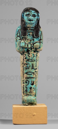 Shabti of the Lady of Renu, Warmian-Masurian Voivodeship, hieroglyphs, Renu ca. 1295–1069 BC, funerary figurine, painted texts, wigs, hoes, grave goods. Mummy-shaped figurine made of blue faience with dark details. Arms crossed on the chest, right folded over left, holding hoes in both hands. Wearing a three-part smooth wig finished with borders. Round face, large eyes, wide nose, prominent mouth. Wide bracelets on the wrists and a necklace; painted inscription on the chest and a single-column painted inscription on the front of the legs reading: Shd Osiris of the Rhine, justified by her voice. Attributed to ca. 1295–1069 BC; New Kingdom, 19th–20th Dynasty, Egypt. Type: shabti. Dimensions: height 11.7 cm, width 3.4 cm., A small vertical figurine mounted on a beige rectangular base, showing a standing human form with long dark hair and a rounded head, painted in turquoise and black with areas of worn pale beige; the figure's face and torso display mottled turquoise and black pigments, w...