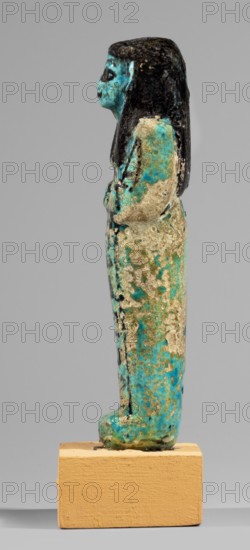 Shabti of the Lady of Renu, Mummy-shaped figurine made of blue faience with dark painted details; arms crossed on the chest right over left holding hoes; three-part smooth wig finished with borders; round face, large eyes, wide nose, prominent mouth; wide bracelets and a necklace. Painted inscription in a single column on the front of the legs reading Shd Osiris of the Rhine, justified by her voice. Ca. 1295–1069 BC; New Kingdom, 19th–20th Dynasty, Egypt. Shabti; height 11.7 cm, width 3.4 cm., A slender standing figure with a long dark hairpiece and profile view, rendered in a mottled mix of turquoise blue, green, and pale beige with areas of dark blackened surface, mounted on a rectangular tan block base.