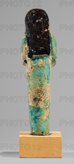 Shabti of the Lady of Renu, Warmian-Masurian Voivodeship, hieroglyphs, Renu ca. 1295–1069 BC, funerary figurine, painted texts, wigs, hoes, grave goods. Mummy-shaped figurine made of blue faience with dark details. Arms crossed on the chest, right arm folded over left, holding hoes in both hands. Three-part smooth wig finished with borders. Round face, large eyes, wide nose, prominent mouth. Wide bracelets on the wrists; necklace with inscription on the chest. Painted inscription arranged in a single column on the front of the figurine's legs: Shd Osiris of the Rhine, justified by her voice. Ca. 1295–1069 BC; New Kingdom, 19th–20th Dynasty, shabti; height 11.7 cm, width 3.4 cm., A vertical figurine-like object mounted on a rectangular block, shown from the back; the top portion is dark black with a rough, mottled surface, the middle and lower shaft areas show a weathered mix of turquoise blue, green, and beige with patchy areas of wear and small perforations; the base block is a warm t...