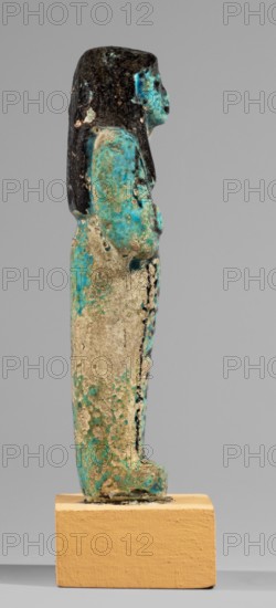 Shabti of the Lady of Renu, Mummy-shaped funerary figurine made of blue faience with dark details. Arms crossed on the chest, right over left, holding hoes in both hands. Wears a smooth three-part wig finished with borders; round face, large eyes, wide nose, prominent mouth. Wide bracelets on the wrists and a necklace; painted inscription on the chest and a painted column of text on the front of the legs reading: Shd Osiris of the Rhine, justified by her voice. ca. 1295–1069 BC; New Kingdom, 19th–20th Dynasty, Egypt. Shabti; height 11.7 cm, width 3.4 cm., A weathered figurine stands upright on a rectangular block, shown in profile facing right with a straight posture and arms close to the body; the figure has a dark hair area contrasting with a body mottled in shades of turquoise, aqua, green, beige, and pale brown, with areas of rough texture and small pits and cracks along the side, and the base block is a warm tan or ochre color against a neutral grey background.