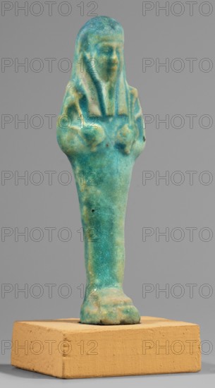 Ushabti; Mummy-shaped funerary figurine standing on a base, supported at the back by a flat pilaster. Arms crossed on the chest, right over left. In the left hand a pickaxe, in the right a hoe and a twisted string from which a basket is slung over the left shoulder. Engraved tools. Long, smooth tripartite wig; oval face with slightly blurred details; long, straight, slightly damaged beard. Not inscribed. Ptolemaic Period, 332–30 BC. From Tuna el-Gebel Hermopolis. Shabti. Height 10.6 cm; width 3.2 cm., A small statuette stands on a rectangular block, showing the upper form of a human figure with a rounded head and a long, narrow body that tapers toward the base; the surface is textured and irregular. The object displays a mix of blue and green hues with areas of pale yellowish and tan discoloration, and there are a few darker spots. The supporting block is a warm beige or light tan color with a rough, slightly pitted surface. The background is a uniform neutral gray.