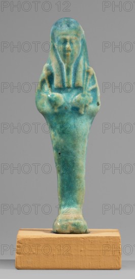Ushabti. Mummy-shaped funerary figurine, standing on a base, supported at the back by a flat pilaster. Arms crossed on the chest, the right folded over the left. In the left hand a pickaxe, in the right a hoe and a twisted string on which a basket is slung over the left shoulder. Engraved tools. On the head a long, smooth, tripartite wig. Oval face with slightly blurred facial details. Long, straight, slightly damaged beard. Not inscribed. Shabti. Ptolemaic Period, 332–30 BC. From Tuna el-Gebel Hermopolis. Height 10.6 cm; width 3.2 cm., A small turquoise and teal-blue glazed figurine with greenish and pale beige weathered areas, showing a standing human-like form with a head covering and arms crossed over the chest, mounted on a rectangular tan-brown wooden block against a plain gray background.