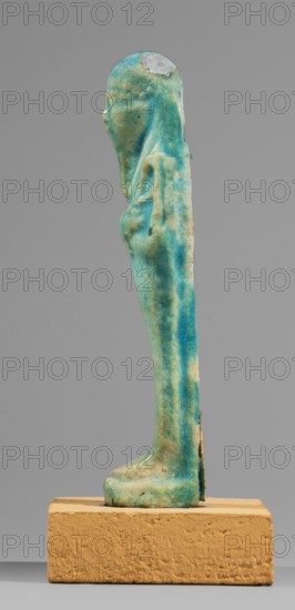 Ushabti, back pillars, hoes, funerary figurine, wig, grave goods, mummy-shaped figurine standing on a base, supported at the back by a flat pilaster. Arms crossed on the chest, the right folded over the left. In the left hand a pickaxe, in the right a hoe and a twisted string on which a basket is slung over the left shoulder. Engraved tools. On the head is a long, smooth, tripartite wig. Oval face, facial details slightly blurred. Long, straight, slightly damaged beard. Not inscribed. 332–30 BC; Ptolemaic Period. From Tuna el-Gebel Tuna el-Galat according to owner, Hermopolis. Shabti. Height 10.6 cm, width 3.2 cm., A narrow upright figurine with a rounded head and stylized features stands on a rectangular block, the figure showing a smooth elongated form with arms held close to the body and simplified feet; the surface of the figurine displays a mottled turquoise and green glaze with areas of lighter cream and small darker spots, and the rectangular base is a warm ochre color.