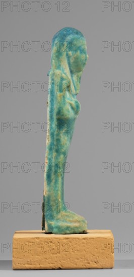 Ushabti, mummy-shaped funerary figurine standing on a base, supported at the back by a flat pilaster. Arms crossed on the chest, right folded over left. Left hand holds a pickaxe, right hand a hoe and a twisted string with a basket slung over the left shoulder. Engraved tools. Wears a long, smooth, tripartite wig. Oval face with slightly blurred facial details; long, straight, slightly damaged beard. Not inscribed. Ptolemaic Period, 332–30 BC. From Tuna el-Gebel Hermopolis. Shabti; height 10.6 cm, width 3.2 cm., A slender vertical statuette in shades of blue and turquoise with areas of greenish and pale yellowish discoloration stands on a rectangular base that is sandy beige in color; the statuette has a rounded head and an elongated body with subtle raised contours suggesting facial profile and torso, and the base appears textured and slightly rough while the background is a flat neutral gray.
