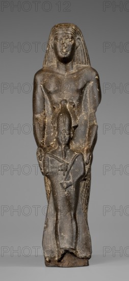 Figurine of a priest with a statuette of the god Osiris. The priest is shown standing, holding in front of himself with both arms a statuette of Osiris, which reaches below his waist. The priest is dressed in a long, pleated loincloth. On his head he wears a wig pushed halfway down his forehead and reaching to his shoulders. The musculature of the torso and arms is clearly defined, with a marked vertical line dividing the torso, and with strongly modeled collarbones. The priest's face is small, modern, with slightly convex, smooth cheeks. Slanted eyes, eyebrows with a gentle arch, artistically rendered. Large ears. Osiris is depicted in a shroud, wearing an atef crown, the front of which is decorated with a uraeus. In his right hand he holds a Nechacha whip, in his left a Heka scepter. The face is round, with large eyes and a slightly smiling mouth. The priest's figure is leaning against a rear pillar reaching to his wig, on which a hieroglyphic inscription is carved; some of the chara...