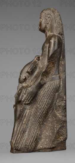 Figurine of a priest with a statuette of the god Osiris, The priest is shown standing, holding in front of himself with both arms a statuette of Osiris, which reaches below his waist. The priest is dressed in a long, pleated loincloth. On his head he wears a wig pushed halfway down his forehead and reaching to his shoulders. The musculature of the torso and arms is clearly defined, with a marked vertical line dividing the torso, and with strongly modeled collarbones. The priest's face is small, modern, with slightly convex, smooth cheeks. Slanted eyes, eyebrows with a gentle arch, artistically rendered. Large ears. Osiris is depicted in a shroud, wearing an atef crown, the front of which is decorated with a uraeus. In his right hand he holds a Nechacha whip, in his left a Heka scepter. The face is round, with large eyes and a slightly smiling mouth. The priest's figure is leaning against a rear pillar reaching to his wig, on which a hieroglyphic inscription is carved; some of the chara...