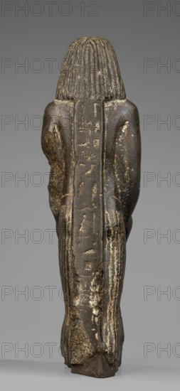 Figurine of a priest with a statuette of the god Osiris. The priest is shown standing, holding in front of himself with both arms a statuette of Osiris, which reaches below his waist. The priest is dressed in a long, pleated loincloth. On his head he wears a wig pushed halfway down his forehead and reaching to his shoulders. The musculature of the torso and arms is clearly defined, with a marked vertical line dividing the torso, and with strongly modeled collarbones. The priest's face is small, modern, with slightly convex, smooth cheeks. Slanted eyes, eyebrows with a gentle arch, artistically rendered. Large ears. Osiris is depicted in a shroud, wearing an atef crown, the front of which is decorated with a uraeus. In his right hand he holds a Nechacha whip, in his left a Heka scepter. The face is round, with large eyes and a slightly smiling mouth. The priest's figure is leaning against a rear pillar reaching to his wig, on which a hieroglyphic inscription is carved; some of the chara...