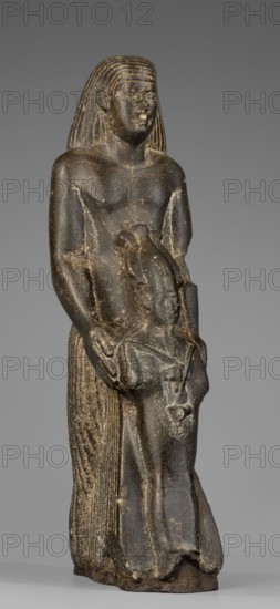 Figurine of a priest holding a statuette of the god Osiris. The priest stands, holding the statuette with both arms; the statuette reaches below his waist. The priest wears a long pleated loincloth and a wig pushed halfway down his forehead to his shoulders. Musculature of torso and arms is clearly defined with a marked vertical line dividing the torso and strongly modeled collarbones. The priest's face is small, modern-looking, with slightly convex smooth cheeks, slanted eyes, gently arched eyebrows, and large ears. Osiris is depicted shrouded, wearing an atef crown fronted by a uraeus; in his right hand he holds a Nechacha whip, in his left a Heka scepter. The priest's figure leans against a rear pillar reaching to his wig, bearing a carved hieroglyphic inscription of which some signs are illegible. Inscribed with hieroglyphs. Late Period, 26th Dynasty, Egypt, 664–525 BC. Sculpture. Height 20.5 cm; width 6 cm; depth 8 cm; weight 0.918 kg., A dark stone statue of a standing figure wit...
