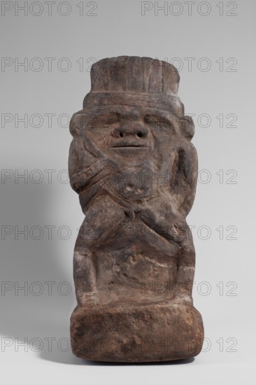 Bes, Bes mythology. Bes naked, standing on a high base, leaning against a pillar. The head is topped with a low crown with four feathers in the shape of triangles widening upwards. The large head and wide face connect directly with the body. The eyes form two depressions occupying a significant part of the face; the nose is wide at the base and widens downwards. The mouth is narrow. On both sides of the face, arched incisions run from the corners of the eyes to the mouth. The body is disproportionately short with a marked navel and a narrow loincloth. The left hand rests on the thigh; the right, very thin, lowered and bent at the elbow, holds a short knife at chest height. The legs are wide apart, excessively tall, thin, and bow-shaped. 3rd century, Edfu; sculpture; height 41.8 cm, width 18.7 cm., A roughly carved stone figure in shades of brown and dark gray stands against a pale gray background; the figure has a broad head with a flattened top, a pronounced brow, recessed eye areas, ...