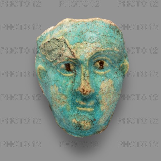 Fragment of a figurine. Oval male face with a protruding chin, small mouth, straight nose, and almond-shaped eyes. Small, protruding ears, forehead receding backward. Unfinished at the back. Egyptian, Ptolemaic Period, 332–30 BC. Terracotta figurine, height 5.9 cm, width 4.5 cm, depth 2.5 cm., A worn sculpted face fragment rendered in turquoise and aqua tones with areas of pale beige and brown weathering, showing a rounded forehead, shallow eye sockets with darker brown spots suggesting pupils, a pronounced nose with brownish discoloration, subtly smiling lips, and textured surface cracks and patches of rougher beige stone against a neutral gray background.