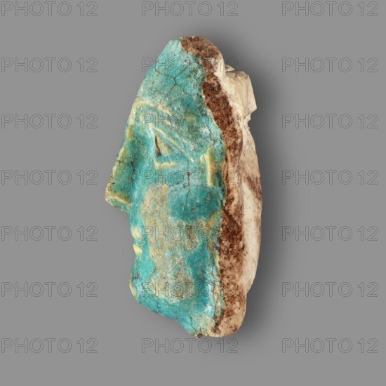 Fragment of a figurine. Oval male face with a protruding chin, small mouth, straight nose, and almond-shaped eyes. Small, protruding ears; forehead receding backward. Unfinished at the back. Egyptian, Ptolemaic Period, 332–30 BC. Figurine, height 5.9 cm, width 4.5 cm, depth 2.5 cm., A side view of a sculpted human face fragment with a turquoise-green glazed surface showing weathered and cracked areas, a few pale yellow highlights on the forehead and near the mouth, and an exposed rough beige and brown stone backing along the rear edge, all set against a uniform gray background.