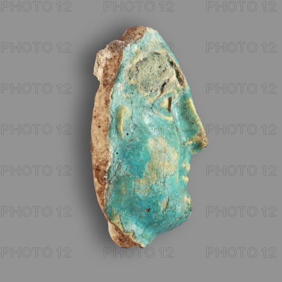 Fragment of a figurine. Oval male face with a protruding chin, small mouth, straight nose, and almond-shaped eyes. Small, protruding ears; forehead receding backward. Unfinished at the back. Egyptian, Ptolemaic Period, 332–30 BC. Figurine; height 5.9 cm, width 4.5 cm, depth 2.5 cm., A worn fragment with a profile-shaped outline showing the contour of a face, displaying a weathered surface of turquoise and aqua tones with areas of pale yellow and muted green; the edges and reverse show beige and brown stone or clay with speckled darker brown spots; the surface has patches of lighter and darker turquoise, small cracks, and irregular rough textures; the background is a flat mid-gray.