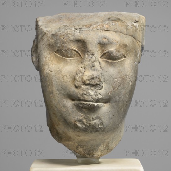 Sculptural model - head of Ptolemy II, Ptolemy II Philadelphus ruler of Egypt; 285–246 BC. Sculpture model, Egyptian sculpture, male head; oval, rather full face, almond-shaped slightly slanting eyes with upper eyelids marked by a double incision. Eyebrows weakly marked, sloping slightly downwards toward the temples. Nose wide; mouth protruding with recessed corners. Ears in the form of two loops. Back of the head smoothed, with a base. Ptolemaic period probably reign of Ptolemy II, Egypt. Height 13.5 cm, width 10.5 cm, depth 9.5 cm, weight 2.59 kg., A weathered stone sculpture of a human head displayed against a gray background, showing a smooth rounded face with a flat top and a band-like element across the forehead, a worn nose and mouth, closed or heavily lidded eyes, and areas of surface erosion and chipping; the sculpture rests on a light-colored stand. Colors visible include gray, beige, tan, and hints of brown.