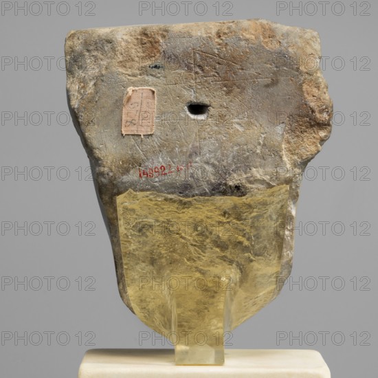 Sculptural model - head of Ptolemy II, Ptolemy II Philadelphus ruler of Egypt; 285–246 BC. Sculpture model of a male head, Egyptian sculpture, oval rather full face, almond-shaped slightly slanting eyes with upper eyelids marked by a double incision. Eyebrows weakly marked, sloping slightly downwards toward the temples. Nose wide; mouth protruding with recessed corners. Ears in the form of two loops. Back of the head smoothed, with a base. Ptolemaic period probably reign of Ptolemy II, Egypt. Height 13.5 cm, width 10.5 cm, depth 9.5 cm, weight 2.59 kg., A roughly rectangular stone fragment with an irregular, weathered surface and a small round hole near its upper center sits mounted on a translucent yellowish support over a pale base; the stone shows colors of gray, beige, brown, and hints of darker spots, and a small rectangular patch of pale fabric is visible on one side.