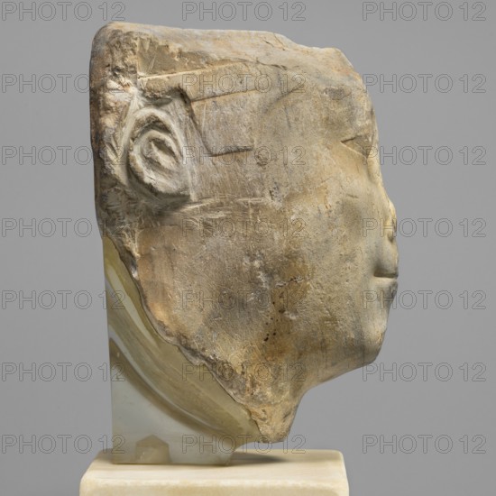 Sculptural model - head of Ptolemy II, Ptolemy II Philadelphus ruler of Egypt; 285–246 BC. Sculptural model, Ptolemaic period probably reign of Ptolemy II, Egypt. Materials/typology: Egyptian sculpture, male head, oval rather full face, almond-shaped slightly slanting eyes with upper eyelids marked by a double incision; weakly marked eyebrows sloping slightly downwards toward the temples; wide nose; protruding mouth with recessed corners; ears in the form of two loops; back of the head smoothed, with a base. Dimensions: height 13.5 cm, width 10.5 cm, depth 9.5 cm, base 19.5 cm, weight 2.59 kg., A weathered stone fragment carved in profile to suggest a face rests on a translucent base and a light platform, showing a rounded forehead, a defined ear with spiral-like inner details, a gently protruding nose, and a subtle closed mouth. The stone surface displays rough, scraped textures and chipped edges, with variations in color including beige, tan, light brown, pale yellow, and hints of gr...