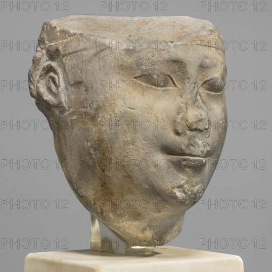 Sculptural model - head of Ptolemy II, Ptolemy II Philadelphus ruler of Egypt; 285–246 BC, sculptural model, Egyptian sculpture, male head, sketches of male heads. Oval, rather full face; almond-shaped, slightly slanting eyes with the upper eyelids marked by a double incision. Eyebrows weakly marked, sloping slightly downwards toward the temples. Nose wide. Mouth protruding with recessed corners. Ears in the form of two loops. Back of the head smoothed, with a base. Ptolemaic period probably reign of Ptolemy II, Egypt. Height 13.5 cm; width 10.5 cm; depth 9.5 cm; weight 2.59 kg., A worn stone sculpture of a human head shown against a neutral gray background, with a smooth blocky top and carved facial features including almond-shaped eyes, a flattened nose, and a faint smile; the left ear is partially visible and the surface shows chips, abrasions, and irregularities. The stone displays shades of gray, beige, tan, and muted brown with subtle darker spots and lighter worn areas. The head...