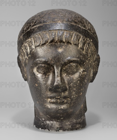 Head of a Ptolemaic prince, unknown creator; diadems; Ptolemy XV Caesarion ruler of Egypt; 47–30 BC probably; kings; Egyptian sculpture; Egyptian art; hairstyles. Head of a man with short hair tied with a headband crossed at the nape of the neck and falling at the ends to the neck. Short, wide strands of hair protrude from under the headband onto the forehead. The face is small, with regular features, a straight nose widening significantly downward, prominent lips, a clearly emphasized chin, large deep-set eyes, prominent eyebrows, small ears close-fitting to the head. The upper fragment of the pilaster is visible at the back, with the head resting on it. With underlay 20.5, 44–30 BC; Ptolemaic period end of the reigns of Caesarion and Cleopatra, Egypt; sculpture. Dimensions: height 19.3 cm, width 13 cm, depth 17 cm; weight 7.532 kg., A sculpted head made of dark stone with lighter worn areas, showing a smooth rounded top, a band around the forehead, and short stylized hair along the h...