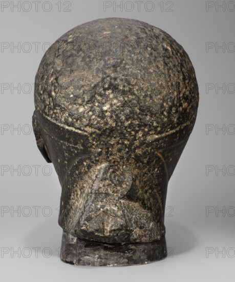 Head of a Ptolemaic prince; diadem; probably Ptolemy XV Caesarion ruler of Egypt; 47–30 BC; Egyptian sculpture, Ptolemaic period end of the reigns of Caesarion and Cleopatra, 20.5, 44–30 BC; limestone, sculpture of a man with short hair tied with a headband crossed at the nape and falling at the ends to the neck. Short, wide strands of hair protrude from under the headband onto the forehead. Small face with regular features, straight nose widening downward, prominent lips, clearly emphasized chin, large deep-set eyes, prominent eyebrows, small ears close to the head. Upper fragment of a pilaster visible at the back, head resting on it. Height 19.3 cm; width 13 cm; depth 17 cm; weight 7.532 kg., The image shows the back of a sculpted head with a rough, pitted surface in dark gray and black tones with lighter gray and tan speckling; the form includes a rounded cranium, a defined nape, and a short, squared base that appears darker and slightly glossy, all set against a smooth, pale gray b...