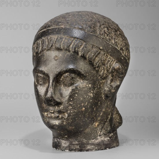 Head of a Ptolemaic prince; diadem; Ptolemy XV Caesarion probably; Egyptian sculpture, Ptolemaic period end of the reigns of Caesarion and Cleopatra, circa 44–30 BC; head of a man with short hair tied with a headband crossed at the nape and falling at the ends to the neck; short, wide strands of hair protrude from under the headband onto the forehead; small face with regular features, straight nose widening significantly downward, prominent lips, clearly emphasized chin, large deep-set eyes, prominent eyebrows, small ears close-fitting to the head; upper fragment of a pilaster visible at the back with the head resting on it; materials: sculpture; dimensions: height 19.3 cm, width 13 cm, depth 17 cm; weight 7.532 kg., A carved stone head of a person shown against a plain light gray background, rendered in dark gray and black tones with patches of lighter gray and brownish weathering; the head has a rounded cap or hairband with textured short hair carved along the forehead and sides, def...
