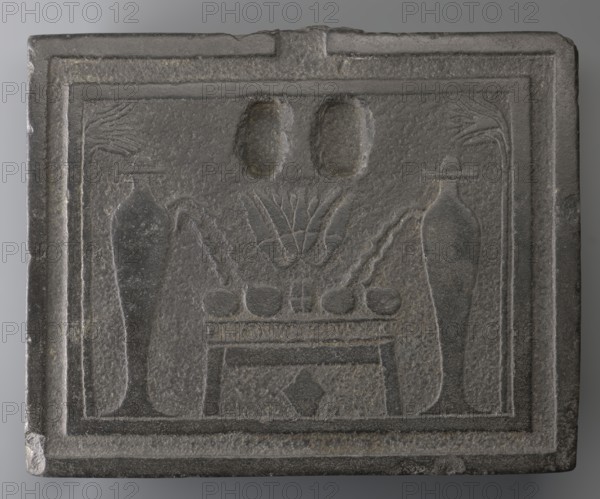 Offering table with carved offerings;, Offering table with offerings depicted: bread and two slender vessels, behind which lotus flowers are visible on high stems. Iconography: offerings, grave goods. Ptolemaic Period, 332–30 BC, Egypt. Dimensions: height 8.5 cm; width 36 cm; depth 29 cm; weight 23.444 kg., A rectangular carved stone plaque in shades of gray with a raised border contains a recessed central panel showing a symmetrical arrangement: a low table or platform with a row of rounded objects on top, flanked by two tall slender vase-like forms with curved handles or spouts, stylized plant motifs rising behind the table, and two oval recessed shapes above; the surface shows a textured, slightly pitted finish and worn edges.