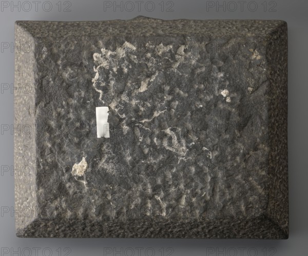 Offering table with carved offerings, offerings iconography and grave goods; depicted: bread and two slender vessels, with lotus flowers on high stems behind; Ptolemaic Period, 332–30 BC, Egypt; offering table; height 8.5 cm, width 36 cm, depth 29 cm, weight 23.444 kg., A rectangular slab with a rough, pitted surface rests against a neutral background; the slab is predominantly dark gray with lighter gray and off-white irregular patches and streaks across its top face, and there is a small bright white rectangular patch adhered near one side. The slab's beveled edges show a mottled texture in shades of gray and muted brown, and subtle shadowing around the slab gives depth against the background.