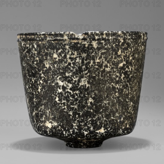 Stone vessel. Vessel with a quadrangular bottom and an almost cylindrical body profiled below the rim. Supported by a small, round foot. Possible Ptolemaic Period, 332–30 BC though it may not be very early — J. Velvet. Egypt. Stone; ritual vessel. Height 8.8 cm, width 10 cm, weight 0.63 kg., A rounded vessel with a slightly flared rim and a small foot, shown against a neutral background; its surface has a speckled, mottled pattern in black and off-white creating a marbled effect, with a small chip at the rim and a subtle shadow beneath the foot; colors visible include black, off-white, gray, and a hint of beige.