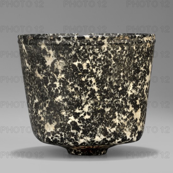 Stone vessel, Königsberg provenance, repossession Frombork; Warmian-Masurian Voivodeship. Ritual stone vessel with a quadrangular bottom and an almost cylindrical body profiled below the rim. The vessel is supported by a small, round foot. 332–30 BC; Ptolemaic Period one should also consider the possibility that this vessel is not very early — J. Velvet. Egypt, vessel. Height 8.8 cm; width 10 cm; weight 0.63 kg., A rounded cup-like object with a slightly flared rim and a small rounded base sits centered against a smooth background, its surface showing a dense mottled pattern of black and white with areas of gray where the two mix; the rim has subtle horizontal ridges and the overall shape tapers gently toward the base, casting a soft shadow beneath.