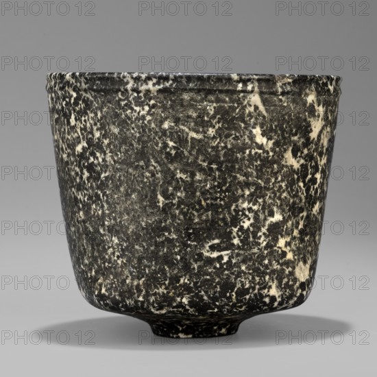 Stone vessel, Königsberg provenance, repossession Frombork; Warmian-Masurian Voivodeship, stone vessel, ritual vessel. Vessel with a quadrangular bottom and an almost cylindrical body profiled below the rim. The vessel is supported by a small, round foot. 332–30 BC; Ptolemaic Period One should also consider the possibility that this vessel is not very early. J. Velvet, Egypt, vessel, height 8.8 cm, width 10 cm, weight 0.63 kg, A close-up view of a rounded cup or vessel with a slightly flared rim, showing a mottled surface of dark gray and off-white patterns that create a marbled appearance; the vessel has a short, slightly raised foot and casts a soft shadow on a smooth gray background.
