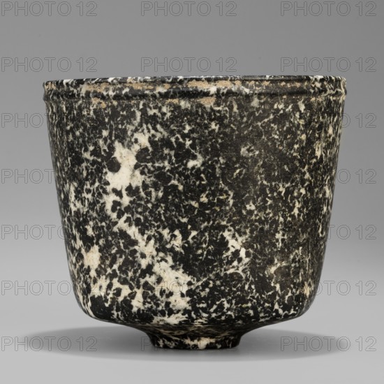 Stone vessel, Königsberg provenance, repossession Frombork; Warmian-Masurian Voivodeship. Stone ritual vessel with a quadrangular bottom and an almost cylindrical body profiled below the rim, supported by a small round foot. 332–30 BC; Ptolemaic Period possibility it is not very early — J. Velvet. Egypt; vessel. Height 8.8 cm, width 10 cm, weight 0.63 kg., A cylindrical cup with a slightly flared rim and a small rounded foot sits against a neutral backdrop; its surface shows a speckled marbled pattern of black and white with a few beige spots, the rim area displaying some worn beige and off-white along the edge, and a soft shadow beneath the cup.