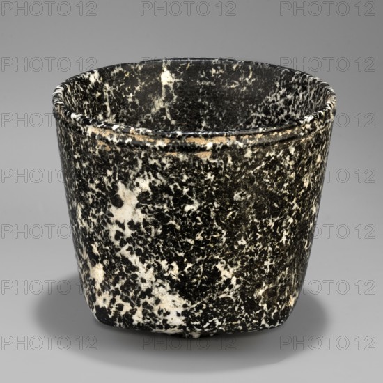 Stone vessel, Königsberg provenance, repossession Frombork; Warmian-Masurian Voivodeship. Stone vessel, ritual vessel. Vessel with a quadrangular bottom and an almost cylindrical body profiled below the rim. The vessel is supported by a small, round foot. 332–30 BC; Ptolemaic Period one should also consider the possibility that this vessel is not very early. Egypt, vessel. Height 8.8 cm, width 10 cm, weight 0.63 kg., A close-up of a rounded cup-like object with a mottled black and white speckled surface, showing a slightly flared rim with a narrow raised band near the top, smooth interior with the same speckled pattern, subtle wear and small lighter patches on the exterior, and a soft gray background casting a faint shadow beneath the object; the visible colors include black, white, gray, and small areas of tan.