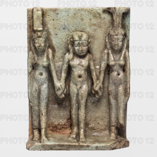 Amulet - plaque with deities: Isis, Nephthys and Horus. Amulet in the shape of a plaque with a triad of deities standing on a base: in the centre Horus, on the left Isis, on the right Nephthys. Back of the plaque is flat. Egyptian deities, hieroglyphs; lock of youth iconography. Late Period, 664–332 BC, Egypt. Amulet plaque, height 4 cm, width 3 cm., A small rectangular relief sculpture showing a group of three standing humanlike figures carved in shallow high-relief and linked by holding hands; the central figure is slightly shorter and more rounded while the two flanking figures are taller and wear tall headpieces, and all three display simplified facial features and visible torsos and limbs. The surface shows a muted pale green color overall with patches of brown and tan staining and some areas of darker discoloration; the material has a slightly glossy, crackled texture and there are small areas where the underlying brownish core is exposed. The base is flat with worn corners and t...