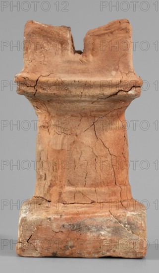 Horned altar serving as a censer, ceremonial vessel/altar, Square base, cylindrical shaft. Lightly profiled Doric base and capital, topped with vertical walls with a single rectangular cutout on each side. 332–30 BC; Ptolemaic Period, Tell Atrib, altar; height 14.5 cm, width 8 cm, weight 0.454 kg., A weathered terracotta object stands against a plain gray background, showing a textured surface with a warm reddish-brown color and areas of lighter orange and pale beige. The piece has a columnar shape with a wider base and a top that features an irregular, notched form; the surface shows many cracks and small chips, rough patches, and uneven edges. There are subtle darker gray and blackened smudges near the base, and traces of white residue in places, while the overall finish appears matte and dusty.