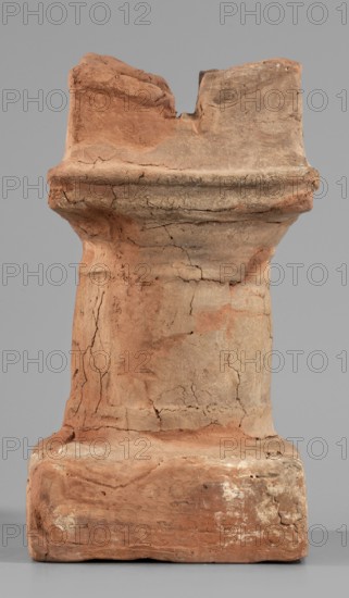 Horned altar serving as a censer, ceremonial vessel/altar. Square base, cylindrical shaft with lightly profiled Doric base and capital, topped with vertical walls with a single rectangular cutout on each side. Ptolemaic Period, 332–30 BC. Found at Tell Atrib. Height 14.5 cm, width 8 cm, weight 0.454 kg., A weathered vertical stone object stands against a plain gray background, showing a rough, cracked surface with a range of earthy colors including reddish-brown, tan, beige, and patches of off-white. The object has a broader base that narrows into a column-like middle and then flares slightly toward the top, with irregular edges and a chipped, uneven top area. Surface texture varies from smooth worn sections to rough crumbly and fractured areas, with visible fissures and pitted spots. Shadows and subtle highlights reveal contours and depth, and traces of lighter residue appear in a few places on the lower portion.