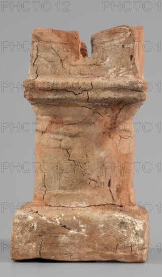 Horned altar serving as a censer, ceremonial vessel/altar with square base and cylindrical shaft; lightly profiled Doric base and capital topped with vertical walls with a single rectangular cutout on each side. Ptolemaic Period, 332–30 BC. Found at Tell Atrib. Altar; height 14.5 cm, width 8 cm, weight 0.454 kg., A weathered, upright clay object with a blocky base narrowing into a cylindrical midsection and flaring slightly at the top, showing many surface cracks and irregularities. The surface displays a mix of reddish-brown, tan, beige, and traces of white residues, with areas of darker brown staining. The top edge is uneven and broken, and the piece has rough, flaky texture and small chips along its contours. The background is a smooth, neutral gray.