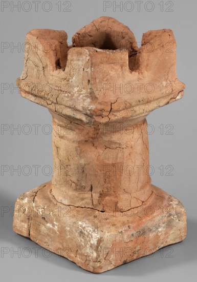 Horned altar serving as a censer, ceremonial vessel/altar with square base and cylindrical shaft, lightly profiled Doric base and capital topped with vertical walls with a single rectangular cutout on each side. Ptolemaic Period, 332–30 BC Tell Atrib, altar; height 14.5 cm, width 8 cm, weight 0.454 kg, A weathered terracotta object in shades of reddish-brown, tan, and beige sits against a neutral gray background, showing a rough, cracked surface with areas of lighter whitish residue; its form includes a square base transitioning into a cylindrical midsection and topped by an irregular, crown-like upper portion with deep indentations and a hollow center, exhibiting visible chip marks, fractures, and textured, uneven edges.