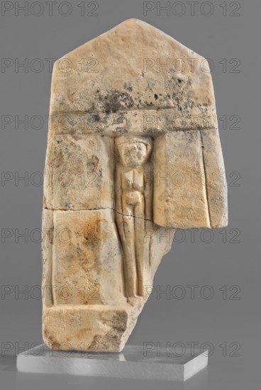 Naos stele with a female figure, small naos-shaped stele slightly tapered at the top and triangular, flat engraving marking the naos design; a female figure schematically represented inside. Stele, height 12 cm, width 6.5 cm., 664–30 BC Late Period–Ptolemaic Period. Found at Tell Atrib., A roughly triangular stone relief fragment in shades of beige, tan, and light brown with darker gray and black spotting stands upright on a translucent base; the carved surface shows a recessed rectangular niche containing a simplified human figure with a rounded head, blocky torso, and long narrow legs, all carved in the same stone color, while the surrounding stone has irregular discoloration, weathering marks, and a crack crossing horizontally; the background behind the object is a uniform medium gray.