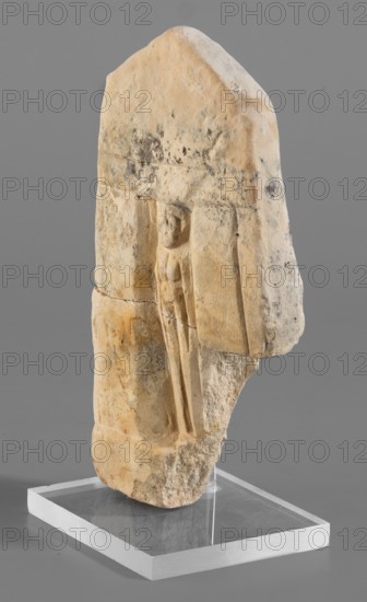 Naos stele with a female figure; small naos-shaped stele, slightly tapered at the top and triangular, with flat engraving marking the naos design. A female figure is schematically represented inside. Stele, height 12 cm, width 6.5 cm., 664–30 BC Late Period–Ptolemaic Period. Found at Tell Atrib., A weathered stone fragment stands upright on a clear rectangular base, showing a carved shallow niche with a small human-like figure in low relief. The stone surface is rough and irregular with worn edges and visible pitting, displaying a palette of beige, tan, cream, and pale yellow with areas of darker gray and black discoloration and some faint orange-brown staining. The carved figure is simplified and elongated, set within vertical recessed panels; the surrounding stone shows tooling marks and natural fractures. The base is transparent and reflective, and the background is a smooth mid-gray.
