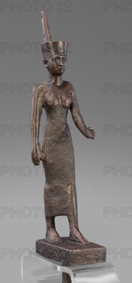Figurine of the goddess Neith. The goddess Neith is depicted in a striding position, with her right arm pressed against the torso and the left bent at the elbow and extended forward. She wears a narrow, tight-fitting dress reaching mid-calf and a red crown with uraeuses. The face is slender and slightly modeled with regular features and very careful modelling. Egyptian art; Egyptian deity. Late Period, 664–332 BC. Terracotta figurine, height 21 cm, width 3 cm, depth 6.2 cm, weight 0.372 kg., A small bronze statue of a standing human figure with a tall cylindrical headdress and an upright staff or plume projecting from it, depicted wearing a long fitted garment that reaches the ankles and standing on a rectangular base; the figure has a smooth stylized face, visible ears, a defined neck with a collar or necklace, one arm extended forward with a slightly cupped hand and the other arm hanging down by the side, with visible feet on the base. The surface shows a rough textured patina with a...