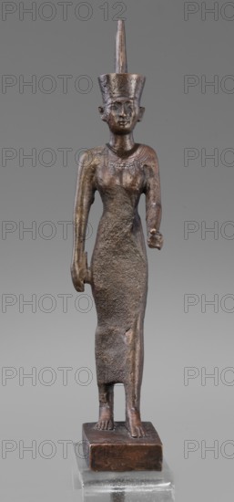Figurine of the goddess Neith, revindication Bytom, Neith mythology, uraeuses, crown iconography, tunic iconography, Scen.Gal.Eg., Egyptian art, Egyptian deities. The goddess Neith is depicted in a striding position, with her right arm pressed against the torso and the left arm bent at the elbow and extended forward. She wears a narrow, tight-fitting dress reaching mid-calf and a red crown on her head. The face is slender and slight, with regular features and very careful modelling. 664–332 BC, Late Period, Egypt. Figurine; height 21 cm, width 3 cm, depth 6.2 cm, weight 0.372 kg., A slender standing statue of a human figure with a tall conical headdress, rendered in a dark brown metallic material with areas of lighter bronze sheen and rougher textured surfaces; the figure faces forward with a stylized face, closed lips, and defined ears, wearing a close-fitting garment that extends to the ankles and shows a textured, worn surface, with both arms down—one hand slightly clenched and the ...