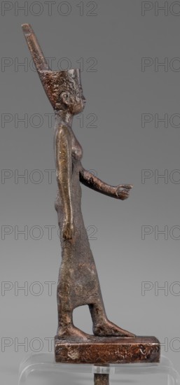 Figurine of the goddess Neith. The goddess Neith is depicted in a striding position, with her right arm pressed against the torso, the left bent at the elbow and extended forward. She wears a narrow, tight-fitting dress reaching mid-calf and a red crown with uraeuses. The face is slender, slight, with regular features and very careful modelling. Egyptian, Late Period, 664–332 BC. Terracotta figurine, height 21 cm, width 3 cm, depth 6.2 cm, weight 0.372 kg., A small bronze figure of a standing person shown in profile with a tall, conical headdress, visible facial features in profile, a slender torso, one arm extended forward with a hand pointing and the other arm hanging down, a long skirt-like lower garment with a cutout near the feet, flat bare feet on a rectangular base, and a smooth neutral gray background; the visible colors include shades of brown, dark bronze, and muted gray.