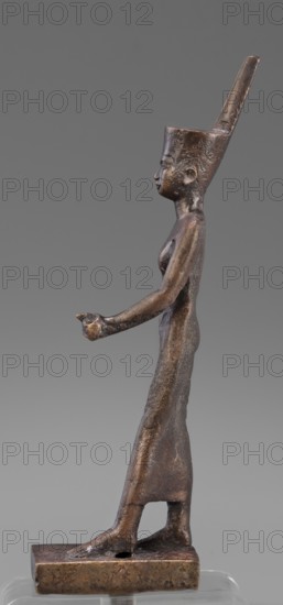 Figurine of the goddess Neith, revindication Bytom, Neith mythology, uraeuses, crown iconography, tunic iconography, Scen.Gal.Eg., Egyptian art, Egyptian deities. The goddess Neith is depicted in a striding position, with her right arm pressed against the torso, the left bent at the elbow and extended forward. She wears a narrow, tight-fitting dress reaching mid-calf, and a red crown on her head. The face is slender, slight, with regular features; very careful modelling. 664–332 BC, Late Period, Egypt. Figurine. Height 21 cm, width 3 cm, depth 6.2 cm, weight 0.372 kg., A slender bronze statuette of a standing human figure shown in profile, with a tall, angular headdress that extends upward and slightly backward, a smooth elongated torso and hips, a long arm extended forward with an open hand, a visible breast, a narrow waist, and legs together ending in bare feet set on a rectangular bronze base; surface shows a textured, aged patina with dark brown and lighter bronze highlights agains...