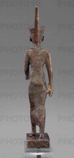 Figurine of the goddess Neith. The goddess Neith is depicted in a striding position, with her right arm pressed against the torso, the left bent at the elbow and extended forward. She wears a narrow, tight-fitting dress reaching mid-calf and a red crown with uraeuses on her head. The face is slender with regular features and very careful modelling. Egyptian, Late Period, 664–332 BC. Material/technique: not specified. Figurine; height 21 cm, width 3 cm, depth 6.2 cm, weight 0.372 kg., A small bronze figurine viewed from behind showing a standing humanlike figure with a tall conical headdress, a slender elongated torso and arms close to the sides, and a long skirt-like lower garment; the surface is textured and pitted, with worn smooth areas, and the piece sits on a square bronze base mounted on a clear rectangular stand; visible colors include brown, dark brown, bronze, and gray, set against a neutral gray background.