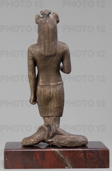 The god Anubis in the form of a jackal-headed deity, Anubis mythology. Figurine of Anubis in the form of a human-jackal-serpent in an upright posture, with the torso slightly tilted back, a jackal head with a strongly elongated muzzle and a mane flowing down the back. Dressed only in an Egyptian apron, below which the torso merges into the body of a snake with a striated abdomen and scaled skin, curled in a horizontal figure eight that supports the figurine. The left arm is held straight alongside the body with a clenched fist; the right is bent at the elbow, with the forearm extended forward. Three protrusions on the head between the ears. Late 1st–2nd century AD; Roman period, Egypt. Terracotta figurine, height 13.5 cm, width 7.1 cm, weight 0.936 kg with base., A bronze-colored statue viewed from the back showing a standing human figure with long textured hair and a flared headdress, wearing a short textured skirt and a fitted upper garment, with one arm bent and the other hanging do...