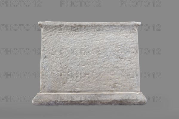 Urn with a depiction of swans, unknown Roman workshop. Sepulchral funerary urn, Roman, dated to the 2nd half of the 1st century AD probably 3rd quarter of the 1st century AD; broadly 2nd half of 1st – 1st half of 2nd century AD. Main plate: trapezoidal box tapering toward the top, placed on a profiled base. Front decorated sparsely; main surface occupied by a tabula ansata with palmette-decorated ears. Below the inscription field are two swans in deep relief, facing each other with strongly arched necks and outstretched wings touching the inscription frame. Upper part crowned by a profiled cornice; on the inner upper edge a rectangular protrusion for securing the lid is visible. Side walls: undecorated; metal pins visible under the cornice on both sides. Lid: original missing. Origin/find: Rome, Roman Empire 27 BC–476. Weight: 45.5 kg., A rectangular stone block with a rough textured front surface sits on a matching base against a plain background. The stone appears mostly light gray w...