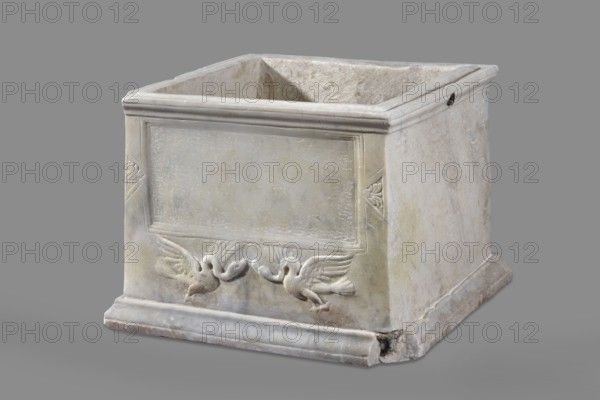 Urn with a depiction of swans. Main plate: the urn's box is trapezoidal, tapering toward the top, placed on a profiled base. The front surface is sparsely decorated; the main area features a tabula ansata whose ears are decorated with palmettes. Below the inscription field are two swans in deep relief, facing each other with strongly arched necks and appearing to take flight, their outstretched wings touching the inscription frame. The upper part is crowned by a profiled cornice; on the inner upper edge a rectangular protrusion for securing the lid is visible. Side walls: no decoration; metal pins visible under the cornice on both sides. Lid: original missing. Dated to the 2nd half of the 1st century AD probably 3rd quarter of the 1st century AD based on analogies; broadly 2nd half of 1st–1st half of 2nd century AD. Roman Empire Rome. Sepulchral funerary urn, weight 45.5 kg. Maker: unknown Roman workshop., A square stone box with an open top, showing worn and slightly rough surfaces in...