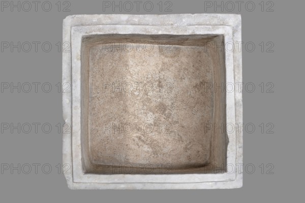 Urn with a depiction of swans, unknown Roman workshop. Sepulchral funerary urn, main plate: trapezoidal box tapering toward the top, placed on a profiled base. Front decoration: a tabula ansata with palmette-decorated ears; below the inscription field are two swans in deep relief, facing each other with strongly arched necks, appearing to take flight with outstretched wings touching the inscription frame. Upper part crowned by a profiled cornice; rectangular protrusion on the inner upper edge for securing a lid. Side walls: no decoration; metal pins visible under the cornice on both sides. Lid: original missing. Dated to the 2nd half of the 1st century AD probably 3rd quarter of the 1st century AD — Roman Empire 27 BC–476 AD. Findplace: Rome. Urn Roman. Weight: 45.5 kg., A top-down view of a square, recessed basin with thick surrounding rim, showing a textured interior surface and visible wear; the basin interior is a mix of beige, tan, and light brown tones with speckled variations, w...