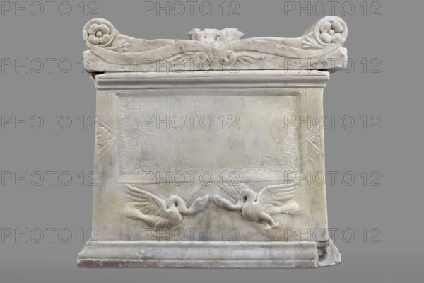 Urn with a depiction of swans, unknown Roman workshop. Sepulchral funerary architecture. Main plate: trapezoidal urn tapering toward the top, placed on a profiled base. Front decoration dominated by a tabula ansata whose ears are decorated with palmettes. Below the inscription field are two swans in deep relief, facing each other with strongly arched necks and outstretched wings touching the inscription frame. Upper part crowned by a profiled cornice; rectangular protrusion on the inner upper edge for securing the lid. Side walls: no decoration. Metal pins visible under the cornice on both sides. Lid: original missing. Dated to the 2nd half of the 1st century AD probably 3rd quarter of the 1st century AD or broadly 2nd half 1st–1st half 2nd century AD. Roman Empire; findspot Rome. Urn Roman. Weight 45.5 kg., A rectangular carved stone monument set against a plain gray background, primarily in shades of off-white, light gray, and pale beige with subtle mottled discoloration; the top edg...